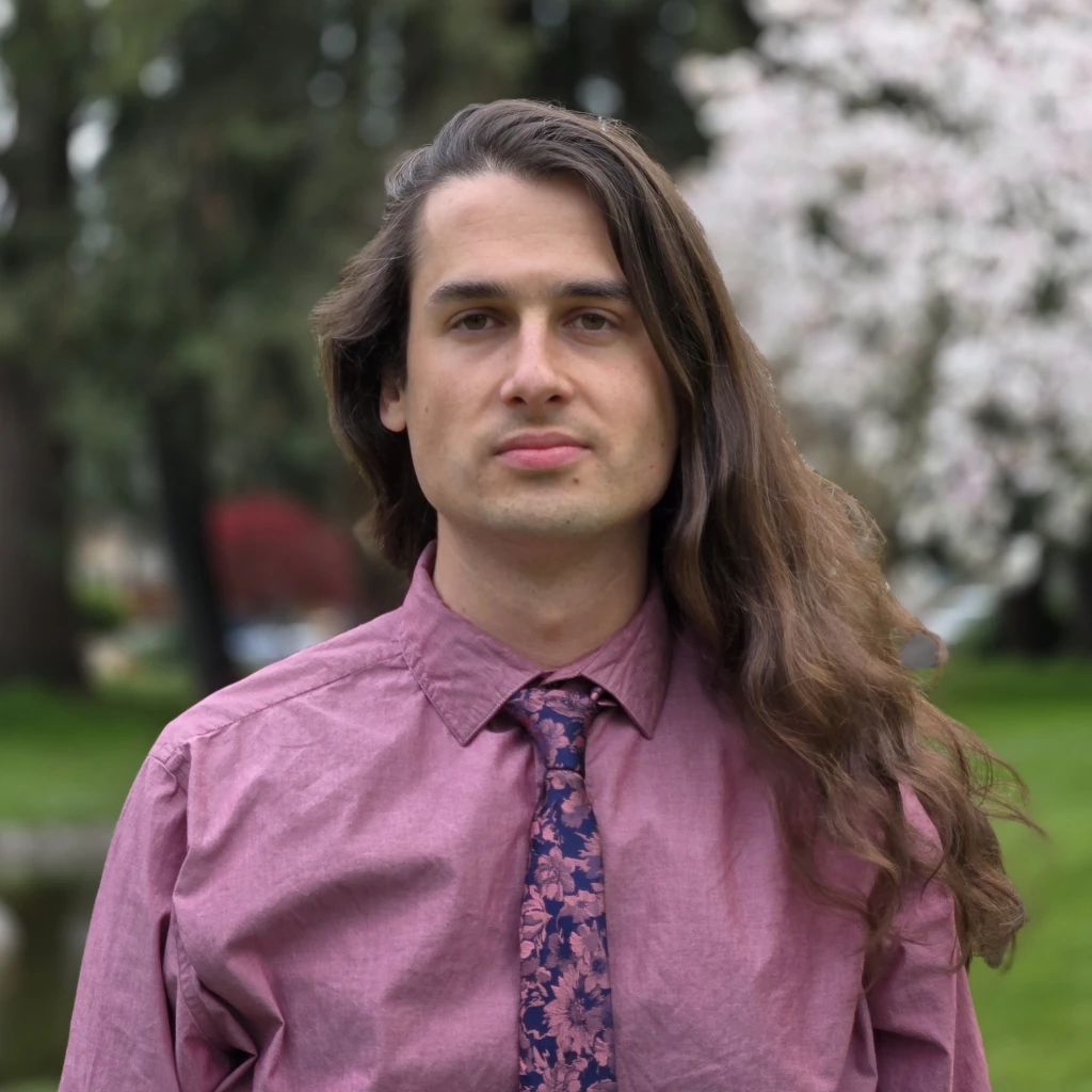 Jay Belmonarcy wearing a light purple shirt with a black and purple tie. His long brown hair is falling on his left shoulder