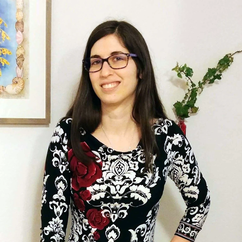 Julie Arnold wearing a dress with white pattern and black background, her black hair is flowing about her shoulders and she is wearing glasses and a smile